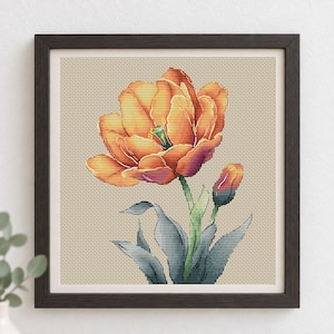 May include: A framed cross-stitch embroidery featuring a large, vibrant orange tulip in full bloom alongside a smaller, unopened tulip bud. The flower has green stems and greyish-blue leaves, all set against a neutral, light beige fabric background.