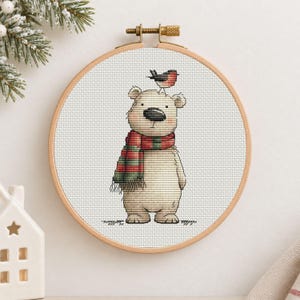 May include: A cross-stitch embroidery in a round wooden hoop featuring a light beige bear wearing a red and green striped scarf with a small bird perched on its head, all stitched onto a white fabric background with a grid pattern.