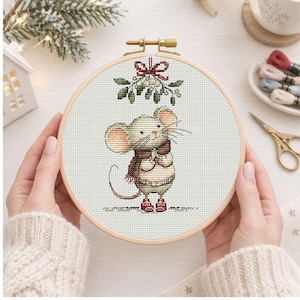 May include: A circular wooden embroidery hoop containing a cross-stitch design of a small mouse wearing a red and green plaid scarf and red boots. Above the mouse is a sprig of mistletoe tied with a red ribbon, all on white fabric.