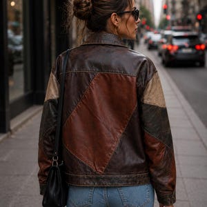 May include: A brown leather jacket featuring a patchwork design with geometric panels in varying shades of dark brown, tan, and reddish-brown, paired with light blue denim jeans and a black shoulder bag with a thin strap.