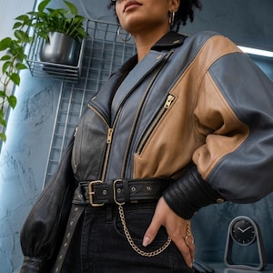 May include: A color-blocked leather jacket featuring panels of black, tan, and slate blue. The jacket includes a notched collar, gold-toned metal zippers, and a wide black leather belt at the waist with a gold-toned buckle and a decorative chain.