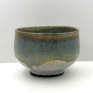 May include: A ceramic bowl featuring a rounded shape and a footed base. The exterior displays a layered glaze effect with a dark brown rim transitioning into a muted blue-green midsection, ending in a white crackle-glazed base with dark lines.
