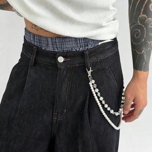 May include: A double-layered wallet chain attached to black denim jeans. One strand features white faux pearls, while the other consists of silver-toned metal beads interspersed with small, colorful sun-shaped charms and clear crystals.
