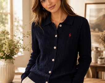 May include: A navy blue long-sleeve cable-knit cardigan featuring a collared neckline, a front button-down closure with light-colored buttons, and a small red embroidered logo on the left chest area, paired with light-colored trousers.