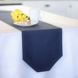 May include: A white tablecloth with a navy blue table runner. A small plate with a pastry and a white mug are on the table runner. A bouquet of yellow flowers is on the table runner.