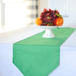 Könnte beinhalten: Ein grüner Tischläufer mit spitzem Ende, der auf einer weißen Tischdecke liegt. Ein weißer Sockel mit einem Strauß roter, orangefarbener und gelber Blumen steht auf dem Läufer.