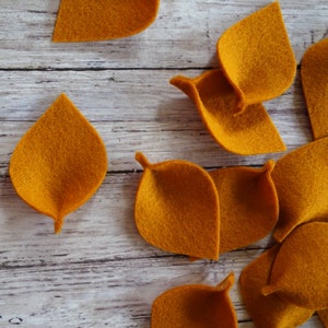 May include: A collection of felt leaf shapes in a burnt orange color. The leaves are arranged on a white wooden surface.