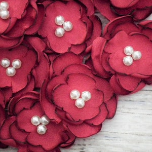 May include: A cluster of burgundy fabric flowers with white pearl embellishments. The flowers are arranged on a white wooden surface.