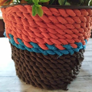 Puede incluir: Una maceta decorativa con un diseño de cuerda trenzada. La maceta presenta bandas de cuerda naranja, turquesa y marrón. La maceta está llena de flores y vegetación artificiales. La maceta es adecuada para uso en interiores o exteriores.