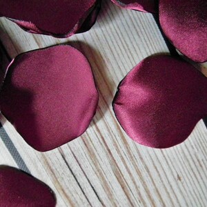 May include: Burgundy satin rose petals with a black edge. The petals are scattered on a wooden surface.