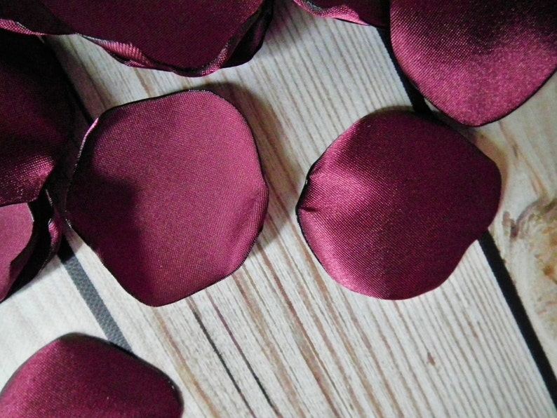 May include: Burgundy satin rose petals with a black edge. The petals are scattered on a wooden surface.