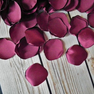 May include: A scattering of burgundy satin rose petals on a wooden surface. The petals are soft and velvety, and they would be perfect for a wedding or other special occasion.