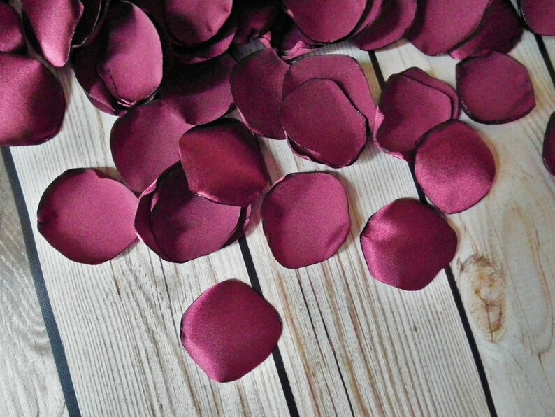 May include: A scattering of burgundy satin rose petals on a wooden surface. The petals are soft and velvety, and they would be perfect for a wedding or other special occasion.