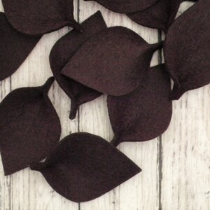 May include: A collection of dark brown felt leaves, arranged in a cluster on a white wooden surface.