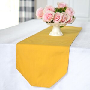 May include: A yellow table runner with a scalloped edge sits on a white tablecloth. A white pedestal holds a bouquet of pink roses.