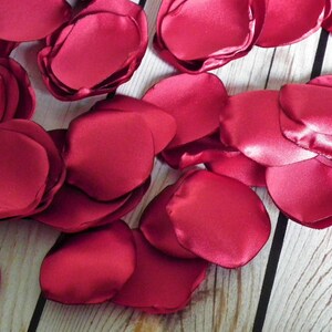 May include: A pile of burgundy satin rose petals, scattered on a wooden surface.
