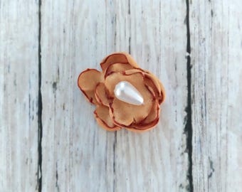 May include: A single peach-colored fabric flower with a white pearl in the center. The flower has a red outline and is on a white wooden background.