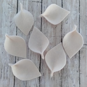 May include: Seven off-white felt leaves with a curved, pointed shape are arranged on a light wood-grain surface. The leaves have a soft, textured appearance and are slightly cupped, suggesting a natural form. These could be used for craft projects.