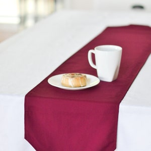 May include: A burgundy table runner with a white plate and a white mug on top of it. The plate has a pastry on it.