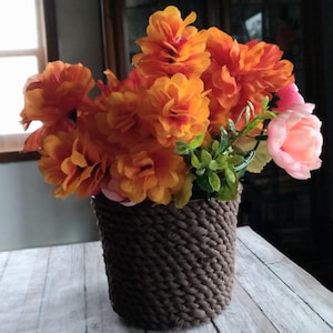 May include: A bouquet of orange and pink artificial flowers in a brown woven pot. The flowers are arranged in a vase on a white wooden table.