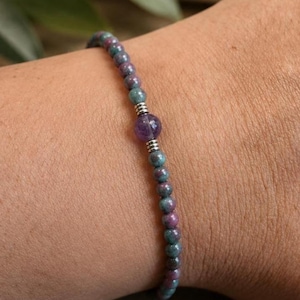 Könnte beinhalten: Ein Perlenarmband mit kleinen, runden, mehrfarbigen Rubin- und Zoisitsteinen in tiefen Grün- und Magentatönen. Das Design umfasst eine einzelne, größere, facettierte violette Amethystperle in der Mitte, die von zwei kleinen, gerippten, silberfarbenen Metallabstandshaltern flankiert wird.