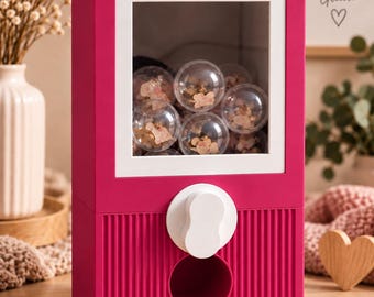 Könnte beinhalten: Ein leuchtend pinker Kaugummiautomat mit einem quadratischen Sichtfenster, das mehrere transparente Kunststoffkugeln mit kleinen Inhalten zeigt. Das Gerät hat einen weißen Drehknopf und eine Ausgabeschale.