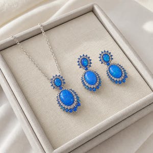 Op de afbeelding: Een sieradenset bestaande uit een ketting met hanger en twee bijpassende oorbellen. Elk sieraad bevat een grote ovale blauwe opaalsteen omringd door een halo van kleinere blauwe en doorzichtige steentjes in een zilverkleurig metalen montuur.