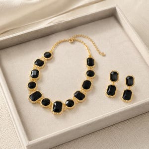 May include: A gold-tone necklace and matching drop earrings set featuring faceted black rectangular stones. The necklace consists of a chain with multiple linked black stones, and the earrings feature two stacked black stones each.