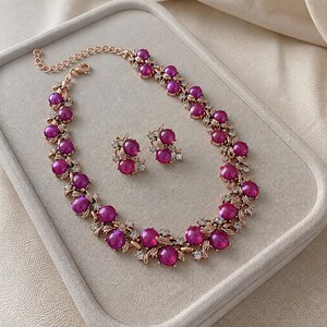 Könnte beinhalten: Ein Schmuckset bestehend aus einer Halskette und passenden Ohrsteckern. Die Stücke zeigen runde, leuchtend magentafarbene Cabochons in einer roségoldfarbenen Metallfassung, verziert mit kleinen, klaren Kristallen und blattartigen Metallelementen.