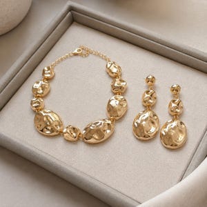Könnte beinhalten: Ein goldfarbenes Schmuckset bestehend aus einem Armband und einem Paar Ohrhänger. Das Armband besteht aus einer Reihe unregelmäßiger, gehämmert wirkender runder Glieder, die durch eine Kette verbunden sind. Die Ohrringe spiegeln dieses Design wider.