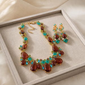 Könnte beinhalten: Ein goldfarbenes Halsketten- und Ohrring-Set mit facettierten tropfenförmigen und quadratischen Kristallen in den Farben Dunkelbraun, leuchtendes Türkis und Limettengrün. Die Halskette verfügt über eine feine Kette mit Karabinerverschluss.