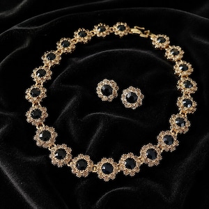 May include: A gold-toned necklace and matching stud earrings set featuring circular flower-shaped links. Each link contains a central round black stone surrounded by a border of small, sparkling clear crystals set in a metallic gold frame.