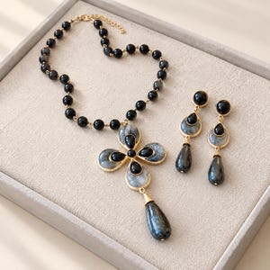 Könnte beinhalten: Ein Schmuckset bestehend aus einer Halskette und passenden Ohrhängern. Die Kette hat schwarze Perlen und einen goldfarbenen Anhänger in Form eines vierblättrigen Kleeblatts mit grauen und schwarzen Steinen. Die Ohrringe haben ein ähnliches Design.