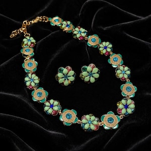 Könnte beinhalten: Ein goldfarbenes Halsketten- und Ohrstecker-Set mit Blumenmotiv. Jede Blüte hat hellgrüne Blütenblätter, einen zentralen blauen Kristall sowie kleine rosa und grüne Schmucksteine. Die Halskette verfügt über eine goldene Kette mit Karabinerverschluss.