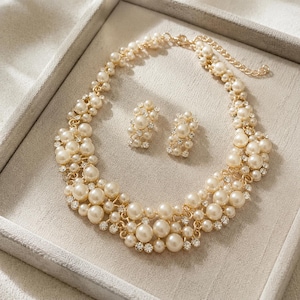 May include: A gold-toned necklace and matching pair of stud earrings featuring clusters of cream-coloured faux pearls and small clear rhinestones. The necklace includes a delicate chain with a lobster clasp and an adjustable extender link at the end.