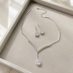 Könnte beinhalten: Ein silberfarbenes Schmuckset bestehend aus einer Halskette und einem Paar passender Ohrringe. Die Halskette hat ein V-förmiges Design mit einem zentralen birnenförmigen, klaren Steinanhänger, während die Ohrringe passende birnenförmige Steine aufweisen.