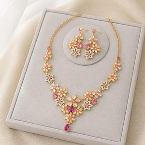 Könnte beinhalten: Ein goldfarbenes Halsketten- und Ohrring-Set mit Blumenmotiven. Der Schmuck ist mit klaren, hellrosa und leuchtend fuchsiafarbenen Kristallen in Blütenform besetzt, wobei ein tropfenförmiger fuchsiafarbener Kristall den Mittelpunkt bildet.
