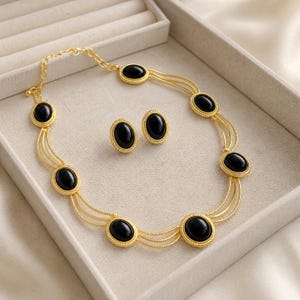 Puede incluir: Un conjunto de collar y pendientes de botón en tono dorado con piedras negras ovaladas tipo cabujón. El collar consta de varias cadenas doradas finas y texturizadas que conectan seis engastes ovalados negros con bordes trenzados.