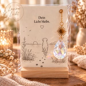 Könnte beinhalten: Eine rechteckige Karte in einer transparenten Kunststoffhülle mit einer schwarzen Strichzeichnung eines Hundes, der auf einen Sonnenuntergang zuläuft, und dem Text "Dein Licht bleibt." Ein goldener Sonnenanhänger und ein facettierter Kristall.