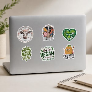 May include: Six vinyl stickers on a laptop lid. One shows a cow with flowers and text, another features various animals with the text LIBERATION ALL, a heart-shaped vegetable sticker, a leaf-shaped sticker, a text sticker, and a potato character.