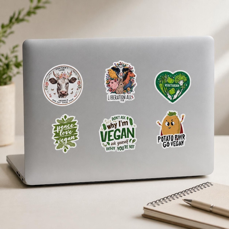 May include: Six vinyl stickers on a laptop lid. One shows a cow with flowers and text, another features various animals with the text LIBERATION ALL, a heart-shaped vegetable sticker, a leaf-shaped sticker, a text sticker, and a potato character.