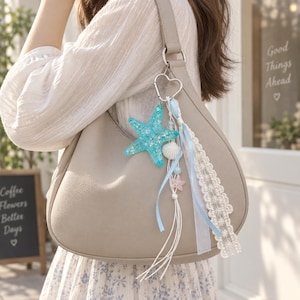 May include: A beige hobo bag featuring a decorative charm attached to the strap. The charm includes a glittery turquoise resin starfish, a small white seashell, a pale pink starfish, light blue ribbons, and a strip of white lace trim.