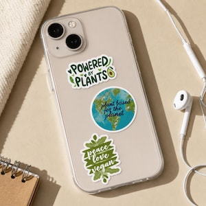 May include: A clear smartphone case decorated with three stickers. The top sticker reads "POWERED BY PLANTS" with an avocado icon. The middle circular sticker shows a globe with the text "plant based for the planet". The bottom sticker says "peace love vegan".