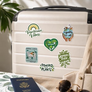 May include: A beige suitcase decorated with five stickers: a green rainbow with 'Vegetarian Vibes', a globe held by hands with 'equality for all', a heart filled with vegetables, a sheep with 'ANIMAL LOVER', and 'POWERED BY PLANTS'.