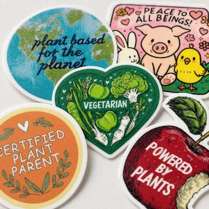 May include: A collection of five stickers featuring various designs: a circular earth with the text plant based for the planet, a rectangular sticker with animals and the text PEACE TO ALL BEINGS, a heart-shaped sticker with vegetables and the text VEGETARIAN, a circular orange sticker with the text CERTIFIED PLANT PARENT, and an apple-shaped sticker with the text POWERED BY PLANTS.