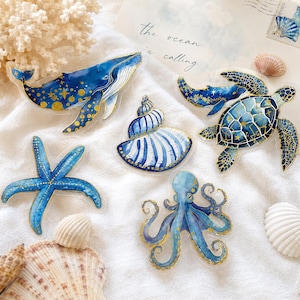 Può includere: Una collezione di cinque ciondoli in acrilico a forma di creature marine, tra cui una balena blu, una tartaruga marina, una stella marina, un polpo e una conchiglia a spirale. Ogni pezzo presenta dettagliati motivi celesti blu e oro.