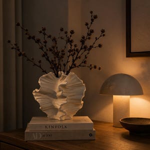 Könnte beinhalten: Eine weiße Keramikvase mit gewellter, muschelartiger Textur, die dunkelbraune Trockenzweige enthält. Die Vase steht auf zwei gestapelten Büchern mit den Titeln Kinfolk und AD at 100, neben einer pilzförmigen Lampe und einer dunklen, flachen Schale.