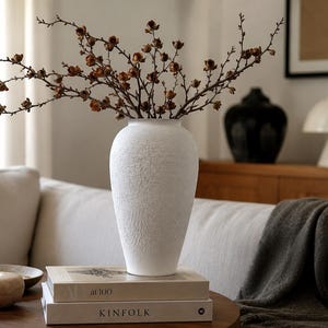 Könnte beinhalten: Eine weiße Vase mit strukturierter, matter Oberfläche, die ein Bündel dünner, brauner, blattloser Zweige mit kleinen, getrockneten Samenkapseln enthält. Die Vase steht auf zwei gestapelten Büchern, eines mit der Aufschrift AD at 100 und das andere KINFOLK.