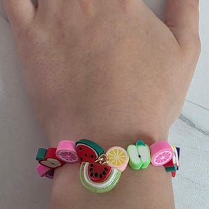 May include: A colorful bracelet featuring various fruit-shaped beads including watermelon slices, citrus segments, and apple shapes. The beads are vibrant shades of pink, green, yellow, and red, strung together to form a playful accessory with a central charm.