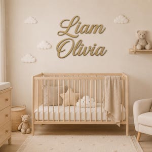 Pode incluir: Dois grandes letreiros de parede em madeira, de cor dourada e em letra cursiva, com os nomes Liam e Olivia, montados numa parede acima de um berço de madeira clara. A parede está decorada com vários pequenos adornos em forma de nuvem.